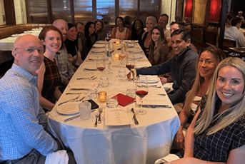A group of people smile for a photo while seated at a long dinner table in a modern restaurant.