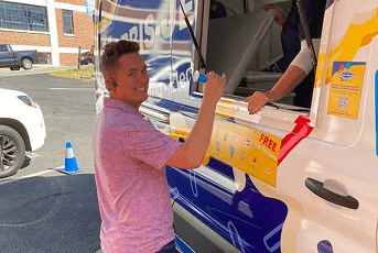 Smiling man in a pink shirt receives a blue ice cream treat from a woman at an ice cream truck window.