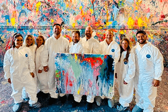 Nine people in white suits pose with a canvas in front of a large, colorful splatter-paint wall.