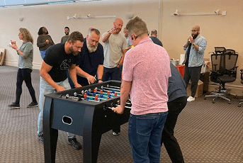 Men playing foosball in a large indoor room as others socialize nearby.